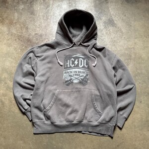 May include: Gray hoodie with a front pocket and a drawstring hood. The front features a graphic of the band AC/DC with the text "BACK IN BLACK 1980" and two guitars. The hoodie is made of a soft, worn material.