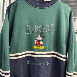Puede incluir: Sudadera verde y azul marino con un dise&ntilde;o bordado de Mickey Mouse. La palabra "Mickey" est&aacute; en el pecho con una fuente vintage. La parte inferior de la sudadera tiene el texto "Established 1928".