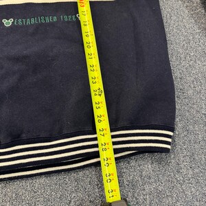 Puede incluir: Sudadera azul marino con rayas blancas en el cuello y los pu&ntilde;os. El texto "ESTABLISHED 1928" est&aacute; impreso en verde. La longitud de la sudadera se mide con una cinta m&eacute;trica amarilla, aproximadamente 76 cm.