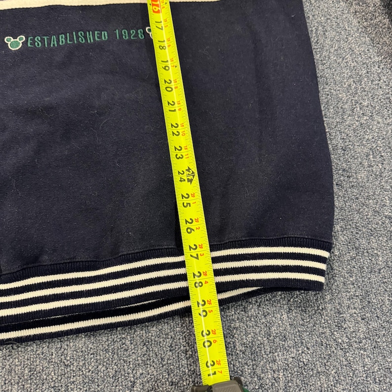 Puede incluir: Sudadera azul marino con rayas blancas en el cuello y los pu&ntilde;os. El texto "ESTABLISHED 1928" est&aacute; impreso en verde. La longitud de la sudadera se mide con una cinta m&eacute;trica amarilla, aproximadamente 76 cm.