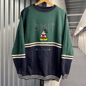 Puede incluir: Sudadera vintage de Mickey Mouse en verde y azul marino. La palabra "Mickey" est&aacute; bordada en el pecho, encima de un dibujo de Mickey Mouse. La parte inferior de la sudadera tiene el texto "Established 1928" y rayas blancas.