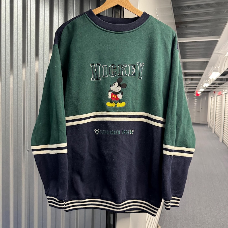 Puede incluir: Sudadera vintage de Mickey Mouse en verde y azul marino. La palabra "Mickey" est&aacute; bordada en el pecho, encima de un dibujo de Mickey Mouse. La parte inferior de la sudadera tiene el texto "Established 1928" y rayas blancas.