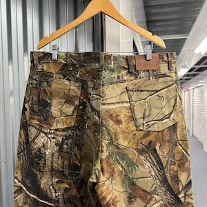 May include: Camouflage-patterned pants hanging on a wooden hanger. The pants feature a brown leather patch with the brand name. The camouflage design incorporates shades of brown, green, and tan, creating a realistic forest-like pattern.