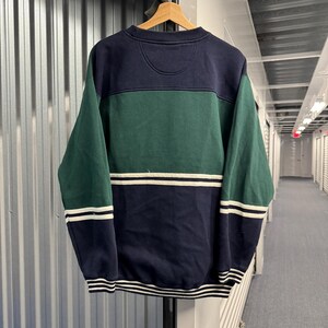 Puede incluir: Una sudadera con bloques de color en azul marino, verde y blanco. La secci&oacute;n superior es azul marino, seguida de una banda verde y una secci&oacute;n inferior azul marino con rayas blancas. Las mangas son verdes con rayas blancas en los pu&ntilde;os.