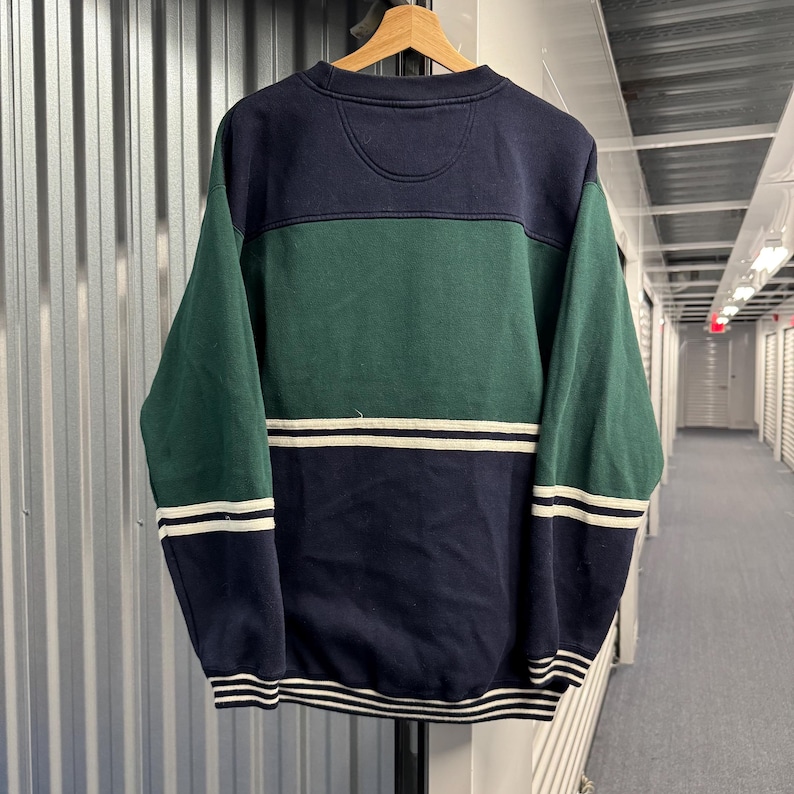 Puede incluir: Una sudadera con bloques de color en azul marino, verde y blanco. La secci&oacute;n superior es azul marino, seguida de una banda verde y una secci&oacute;n inferior azul marino con rayas blancas. Las mangas son verdes con rayas blancas en los pu&ntilde;os.