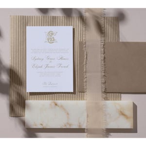 May include: A flat lay featuring wedding stationery. A white invitation with gold lettering and a monogram is centered, with a kraft paper envelope, a marble tile, and a ribbon. The invitation reads "Sydney Grace Klaus & Elijah James French".