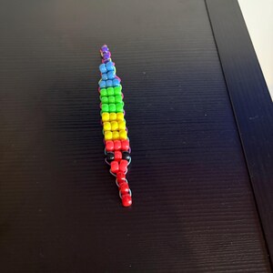 May include: A beaded keychain in a rainbow color scheme. The keychain is made of small, square beads in shades of red, orange, yellow, green, blue, and purple. The beads are strung together to form a long, slender shape.