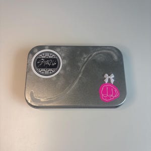 May include: A rectangular, silver-colored tin with a decorative design. The tin features a circular sticker with black text and a pink fist graphic with a white bow. The tin is on a white surface.