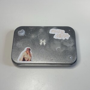 Könnte beinhalten: Eine rechteckige, silberfarbene Metalldose, die mit Aufklebern verziert ist. Zu den Aufklebern gehören ein Herz, eine Schleife und ein Aufkleber mit dem Text "FELIX KARMA". Es gibt auch einen Aufkleber einer Person.