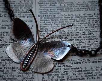 May include: A silver butterfly pendant necklace with a copper chain. The butterfly wings have a hammered texture and iridescent highlights. The pendant is hanging on a page of a book with text about butterflies.