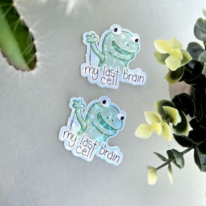 May include: Two stickers featuring a cartoon frog with the text "my last brain cell." The frog is light blue with a holographic effect, waving with a smile. The stickers are on a light gray surface, with green and yellow foliage in the background.