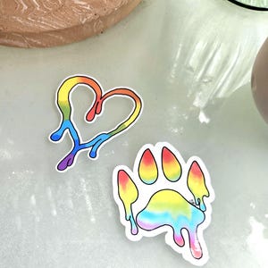 May include: Two stickers: a rainbow-colored heart and a paw print. The heart is outlined in black with a dripping effect. The paw print has a white border and a rainbow gradient with a dripping design. Both stickers are on a light-colored surface.
