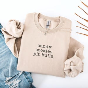 May include: A beige sweatshirt with the words "candy cookies pit bulls" printed in black. A silver necklace with a small pendant is on the sweatshirt. The sweatshirt is folded with a pair of blue jeans.
