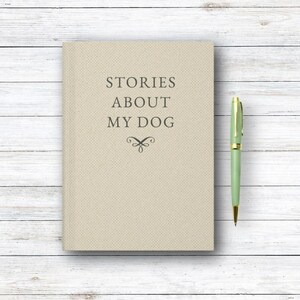 May include: A beige journal with the text "STORIES ABOUT MY DOG" and a decorative flourish. A green and gold pen rests beside the journal on a white wooden surface. The journal is a hardcover book.