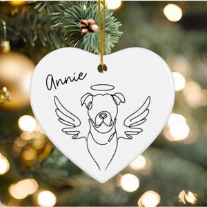 May include: White heart-shaped Christmas ornament with the name "Annie" in black script. A black line drawing of a dog with angel wings and a halo is featured on the ornament. A gold string is attached for hanging.