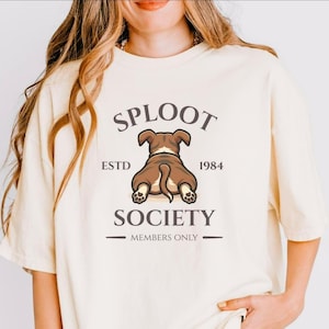 May include: Cream-colored t-shirt featuring a brown dog graphic with the text "SPLOOT SOCIETY ESTD 1984 MEMBERS ONLY." The dog is depicted from the rear, with its tail and paws visible. The shirt has a relaxed fit.