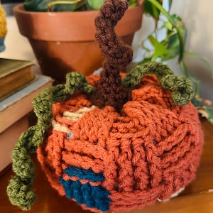 Op de afbeelding: Een gehaakte pompoendecoratie met een textuurrijk oranje lichaam, een donkerbruine krullende steel en groene krullende ranken. De pompoen bevat kleine stukjes crèmekleurig en donkerblauw garen verwerkt in het oranje gehaakte oppervlak.