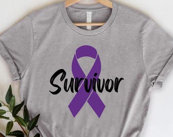 May include: A heather gray t-shirt featuring a purple awareness ribbon and the word "Survivor" in a black script font. The shirt is displayed on a wooden hanger against a white background, with a sprig of greenery in the lower left corner.