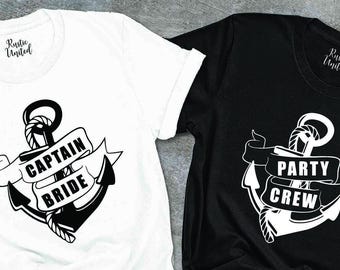 May include: Two t-shirts, one white and one black, featuring nautical anchor designs. The white shirt displays "CAPTAIN BRIDE" on a banner, while the black shirt reads "PARTY CREW." Both shirts have a crew neck and short sleeves.