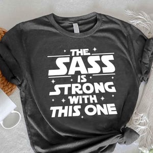 May include: Dark gray t-shirt with white text. The text reads 'THE SASS IS STRONG WITH THIS ONE' in a Star Wars-inspired font. The shirt is on a white surface with a woven placemat and a white mug. A sprig of white flowers is also present.