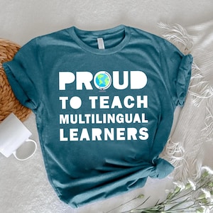 May include: Teal t-shirt with the text "PROUD TO TEACH MULTILINGUAL LEARNERS" in white block letters. The word "PROUD" has a globe graphic. The shirt is knotted at the bottom and is laying on a white surface with a woven mat, a white mug, and flowers.