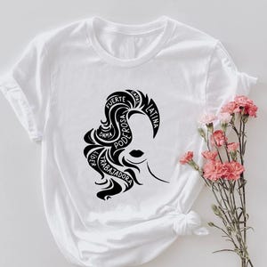 May include: White t-shirt featuring a black graphic of a woman's silhouette with text. The text includes words like "Fuerte", "Poderosa", and "Latina". A bouquet of pink flowers is to the right of the shirt.