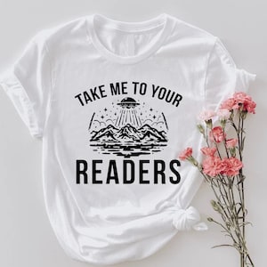 May include: White t-shirt with a black graphic featuring a UFO over mountains, stars, and the text "TAKE ME TO YOUR READERS". The shirt has short sleeves and a round neckline. Pink flowers are on the right side of the shirt.