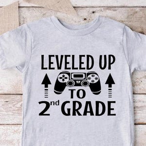 May include: A light gray t-shirt with the text "LEVELED UP TO 2nd GRADE" in black. A black controller graphic is centered above the text. Two black arrows point upwards. The shirt is on a light wood surface.