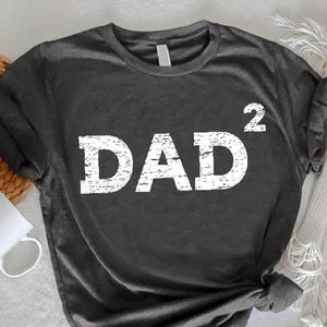 May include: Dark gray t-shirt with the word "DAD²" in distressed white lettering. The shirt is a crew neck style and appears to be made of a soft material. The shirt is knotted at the bottom. A white mug and flowers are in the background.