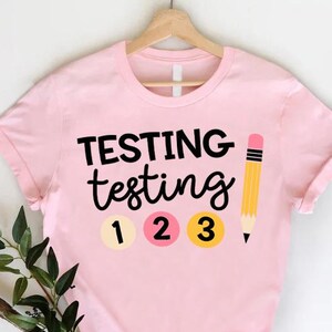 May include: A light pink t-shirt with the words "TESTING testing" in black script. Below the text are the numbers 1, 2, and 3 in circles. A yellow pencil with a pink eraser is to the right of the text. The shirt is hanging on a wooden hanger.