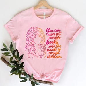 May include: A light pink short-sleeved t-shirt featuring a graphic illustration of a person with voluminous hair and the text, You can never get enough books into the hands of enough children, printed in a gradient of pink, orange, and yellow.
