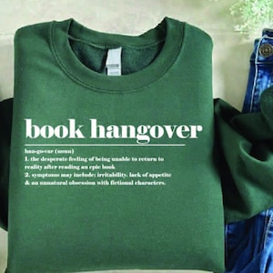 May include: A dark green sweatshirt with the text "book hangover" in white. The definition of "book hangover" is listed below the text. The sweatshirt is paired with blue jeans and a decorative cactus.