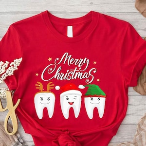 May include: A red t-shirt with the words "Merry Christmas" in white script. The shirt features three cartoon teeth wearing festive hats: reindeer antlers, a Santa hat, and an elf hat. The design is perfect for the holiday season.