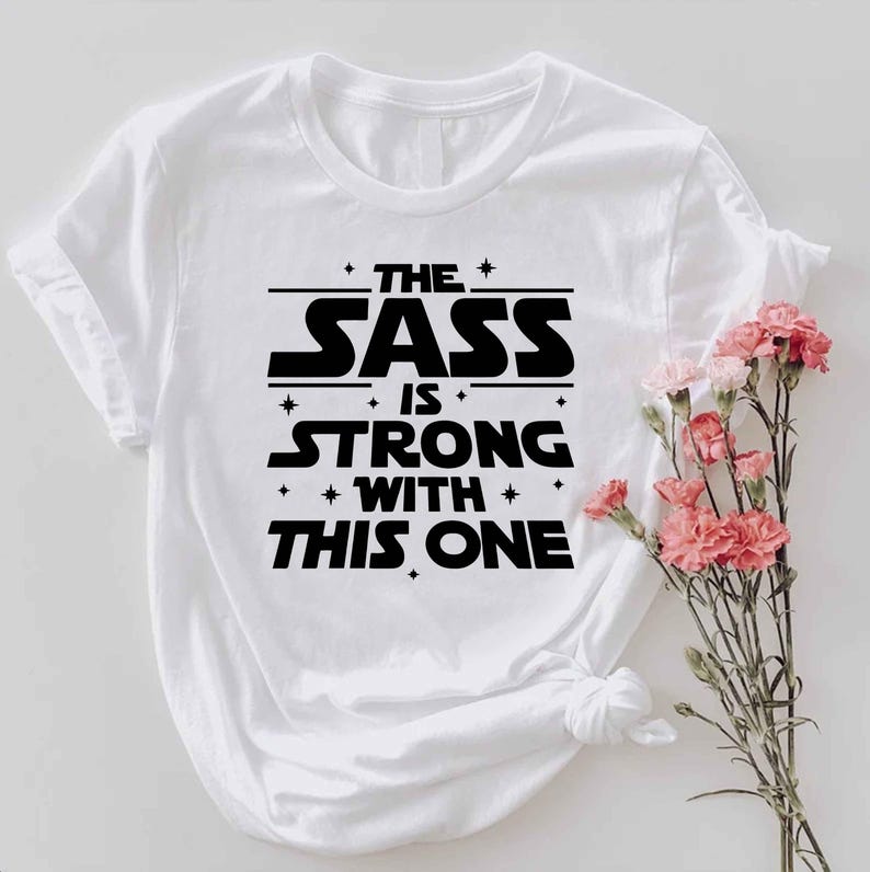 May include: White t-shirt with the text "THE SASS IS STRONG WITH THIS ONE" in black, stylized lettering. The design includes stars and lines. Pink flowers are to the right of the shirt.