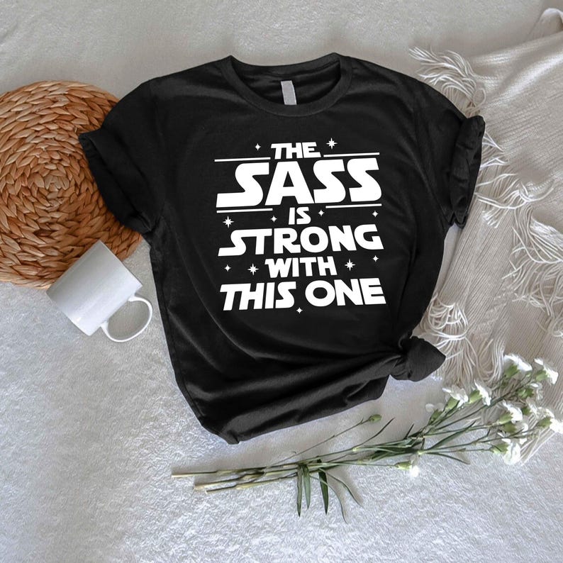 May include: Black t-shirt with the white text "THE SASS IS STRONG WITH THIS ONE." The design features a bold, stylized font with star accents. The shirt is a classic crew neck style.