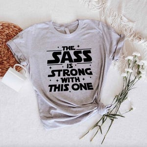 May include: A light gray t-shirt with the text "THE SASS IS STRONG WITH THIS ONE" in black. The shirt is knotted at the bottom right. A white mug and flowers are in the background.