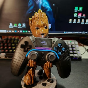 May include: A brown and tan Groot-themed controller stand holding a black controller with blue accents. The stand features a detailed Groot figure with a smiling face and outstretched arms. The controller has a cross, two joysticks, and other buttons.