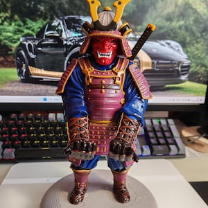 May include: A samurai figurine featuring a red oni mask, a purple and gold chest plate, and blue sleeves. The figure wears a yellow-accented helmet and stands on a circular grey base with a sword hilt visible over the shoulder, arms extended forward.