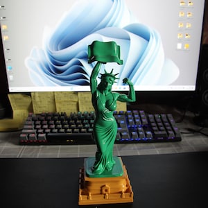 May include: A green figurine of a figure wearing a crown and holding a rectangular object aloft with one arm, while flexing the other arm. The figure stands on a square, orange, tiered base that resembles a pedestal with architectural details.