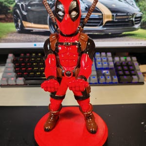 May include: A Deadpool figurine standing on a circular red base, featuring a red and black suit, white eye patches, and two brown sword hilts protruding from the back. The character has outstretched hands, a brown utility belt, and brown boots.