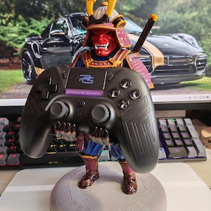 May include: A figurine of a samurai warrior wearing a red mask and traditional armour, standing on a circular grey base. The samurai holds a black gaming controller featuring the text Proove Gaming and a blue logo on its central panel.
