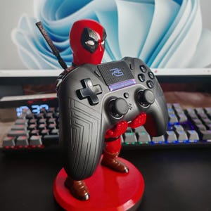 May include: A Deadpool-themed controller stand. The stand features a red base and a Deadpool figure holding a black game controller. The controller has a cross-shaped directional pad and multiple buttons. The stand is designed to hold a game controller.