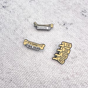 May include: Three enamel pins with medical puns. One pin says "Nailed It" with a bone and hammer. Another says "It's going tibia okay" with a bone. The third says "I've got your back" with a spine.