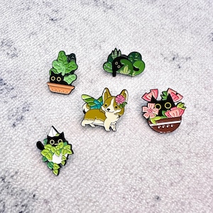 Może przedstawiać: Zestaw emaliowanych przypinek z kotami i corgi. Przypinki przedstawiają czarnego kota w doniczce, corgi ze skrzydłami i kwiatem oraz czarnego kota wśród liści. Przypinki mają błyszczące wykończenie i są na białym tle.
