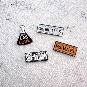 May include: A collection of science-themed pins. One pin features a black and white beaker with the words "LIVE LAB LOVE." Other pins display the word "GENIUS" and "POWER" using elements from the periodic table.
