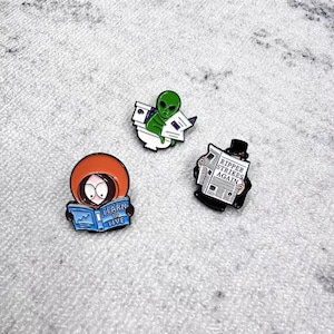 May include: Three enamel pins with cartoon designs. One pin features a character with an orange hood reading a book with the text "LEARN TO LIVE". Another pin shows a green alien reading. The third pin depicts a newspaper with the headline "RIPPER STRIKES AGAIN".