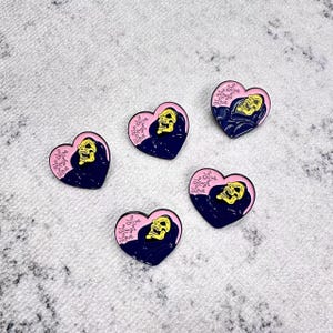 Puede incluir: Cinco pines de esmalte en forma de corazón. Cada pin presenta una calavera amarilla con una capa negra, sobre un corazón azul oscuro. La frase "Live Laugh Love" está escrita en rosa. Los pines están sobre una superficie blanca texturizada.