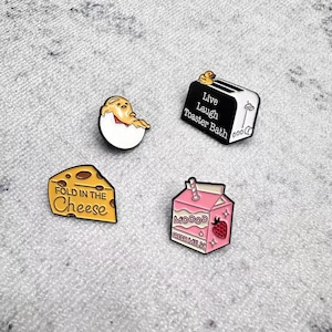 Puede incluir: Cuatro pines de esmalte: un huevo con un pollito, una tostadora en blanco y negro con "Live Laugh Toaster Bath", una cuña de queso con "Fold in the Cheese" y un cartón de leche rosa con una fresa y "Mood Fresh Milk".