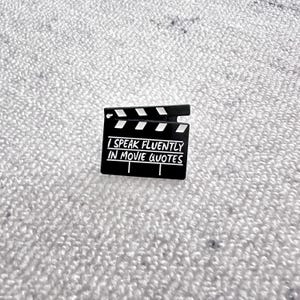 May include: A black enamel pin in the shape of a movie clapperboard. The pin features the text "I SPEAK FLUENTLY IN MOVIE QUOTES" in white lettering. The clapperboard has white and black accents.