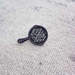 Puede incluir: Un pin de esmalte negro y morado con forma de sartén. El pin presenta el texto "Some of y'all's cornbread ain't done in the middle" en letras blancas. El pin tiene un mango y una superficie de cocción circular.